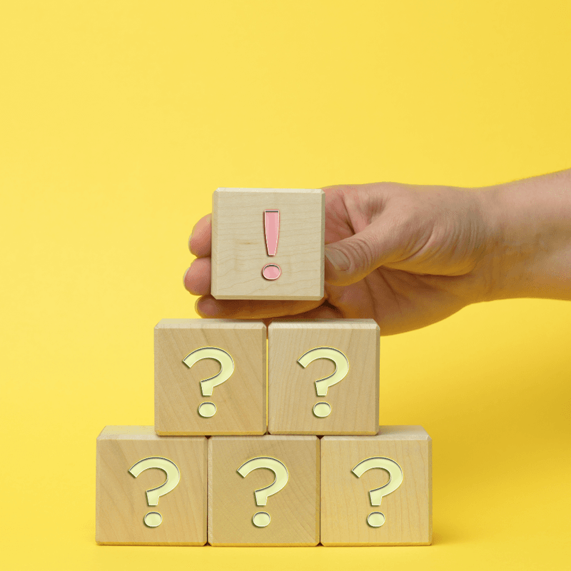 Communicating Compensation Data with Clarity A hand places a small wooden block with an exclamation mark on top of a pyramid of wooden blocks marked with question marks, set against a bright yellow background, symbolizing clarity or answers emerging from uncertainty.